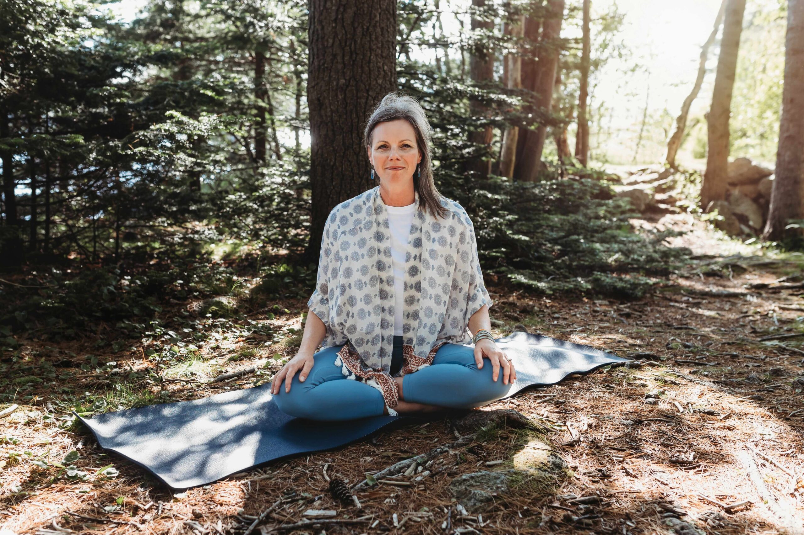 Michelle Robinson (she/her) sitting in a cross-legged pose on a blue yoga mat in a quiet forest setting with sun beams streaming through